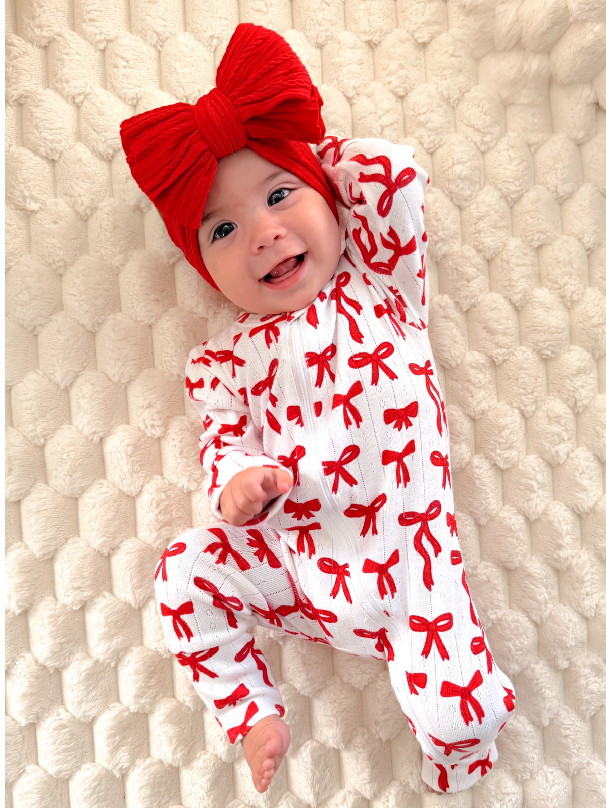 Smiling baby in a red bow headband and patterned onesie on a cozy blanket.