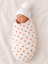 Sleeping newborn wrapped in pumpkin-patterned blanket, wearing a white pom-pom hat on a soft background.