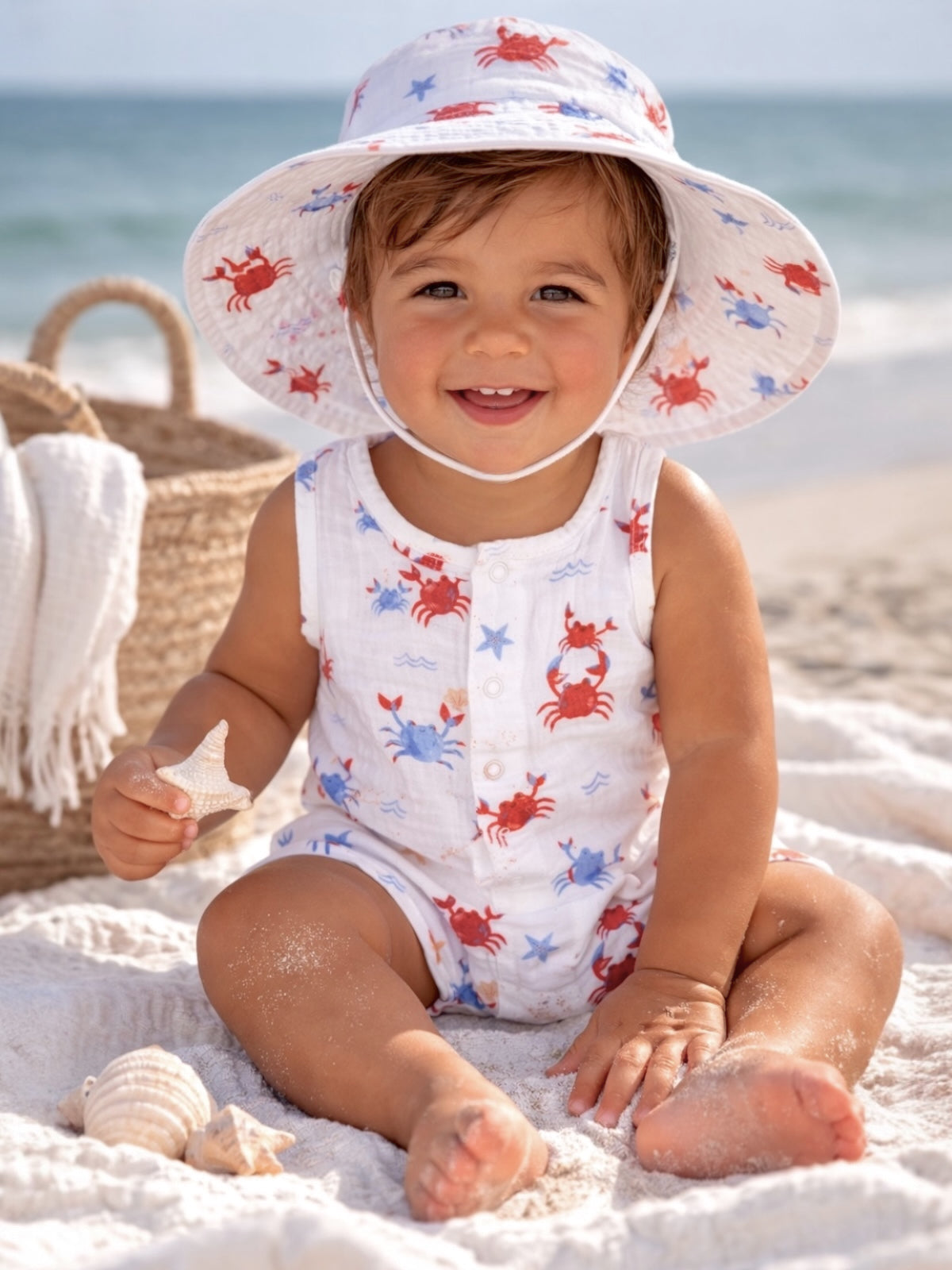 Smiling toddler in a crab-patterned outfit sitting on the sandy beach, holding a starfish.