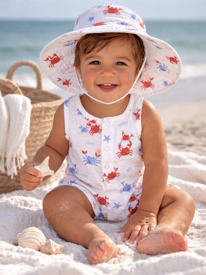 Smiling toddler in a crab-patterned outfit sitting on the sandy beach, holding a starfish.