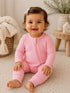 Smiling baby in a pink outfit sitting on a soft rug, surrounded by cozy decor and greenery.