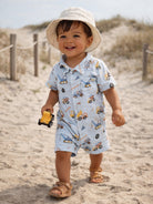 Smiling toddler in a patterned romper, holding a toy truck, walking on a sandy beach with grass in the background.