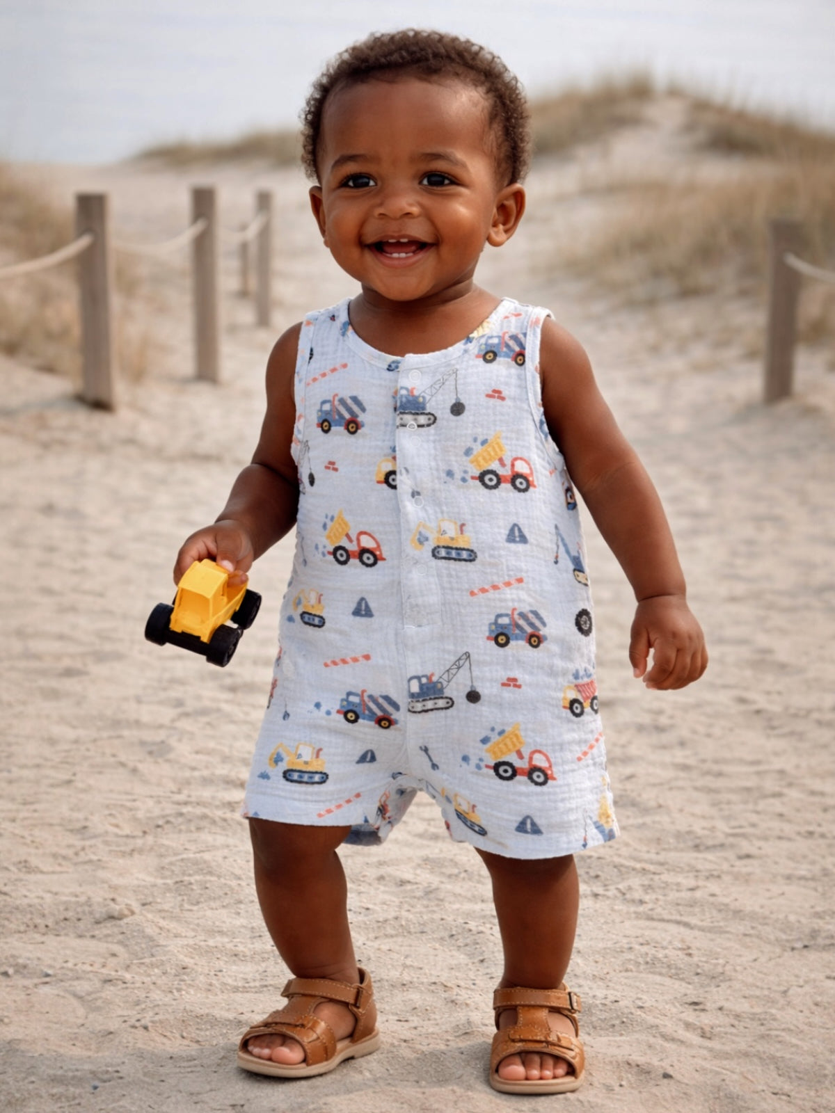 Smiling toddler in a truck-patterned outfit, holding a toy truck on a sandy beach.