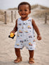 Smiling toddler in a truck-patterned outfit, holding a toy truck on a sandy beach.