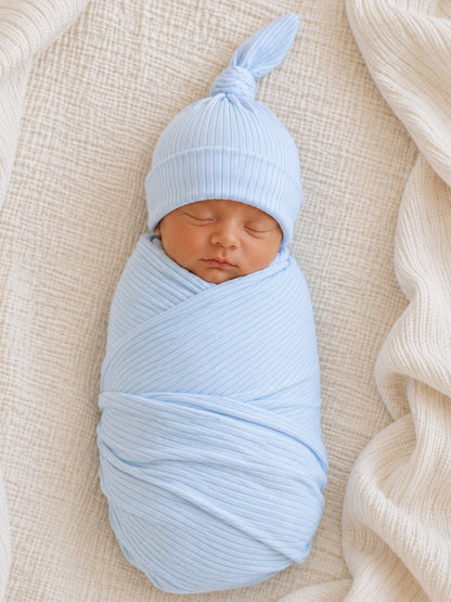 Newborn baby wrapped in light blue fabric, wearing a matching hat, resting on a textured cream blanket.