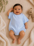 Smiling baby in a blue outfit lying on a textured blanket, next to a teddy bear and greenery.