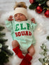 Newborn in an "Elf Squad" outfit with a cream hat and red booties, surrounded by festive greenery and ornaments.