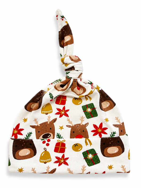 White fabric baby hat with festive reindeer, flowers, and gift prints, tied at the top. Ideal for winter holidays.