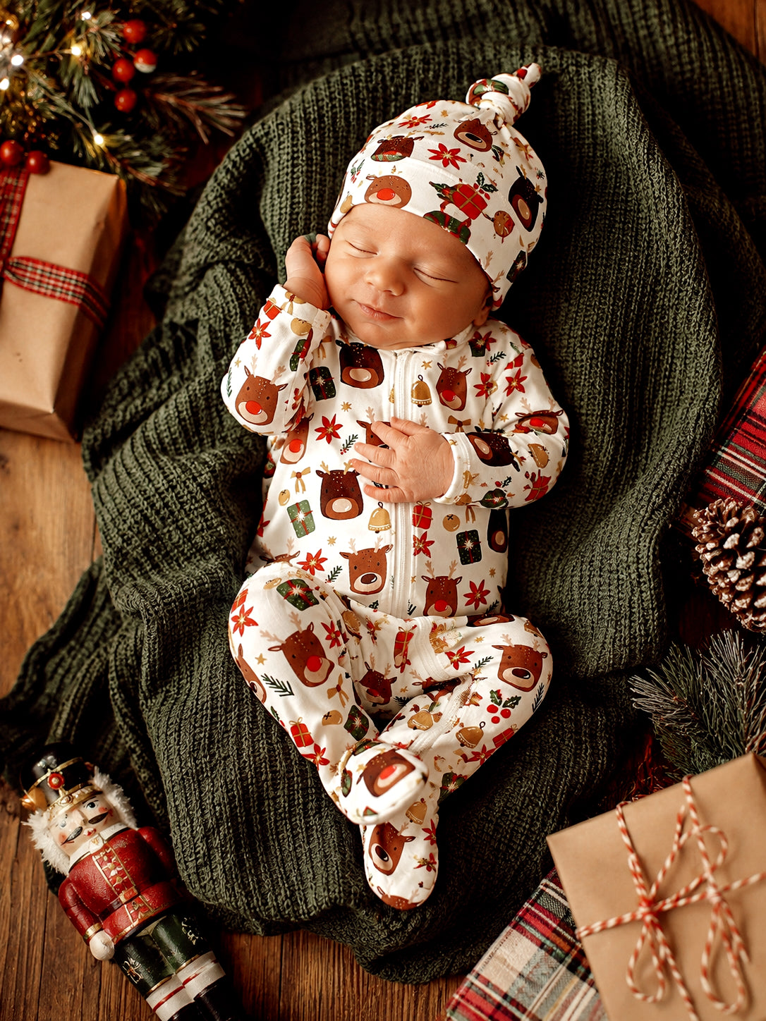 A sleeping baby in festive patterned pajamas, nestled on a cozy green blanket surrounded by holiday decor.