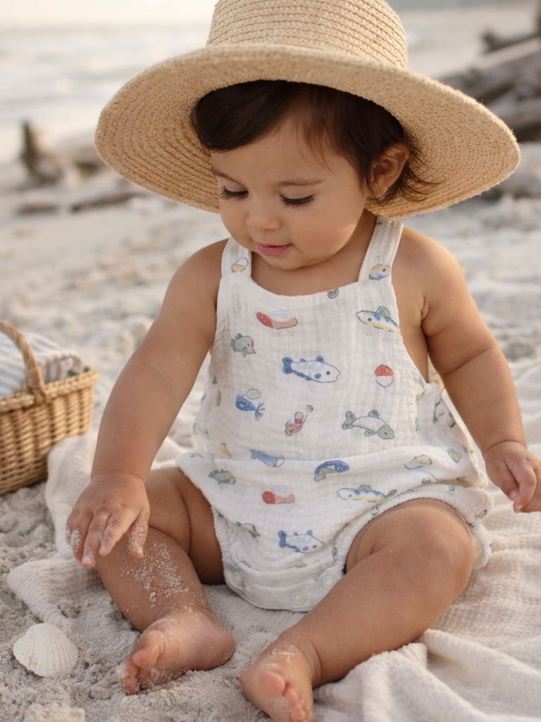 Toddler in a straw hat playing in sand, wearing a colorful fish-patterned outfit at the beach.