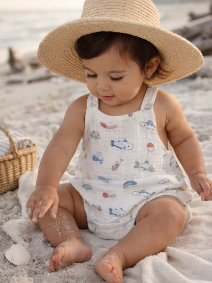 Toddler in a straw hat playing in sand, wearing a colorful fish-patterned outfit at the beach.
