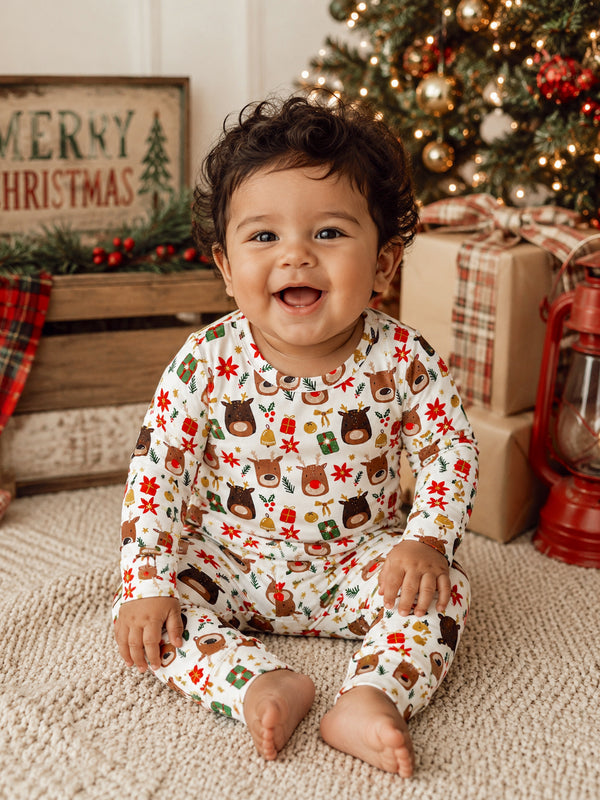 Smiling baby in festive clothing sitting on a rug near a Christmas tree and holiday decorations.