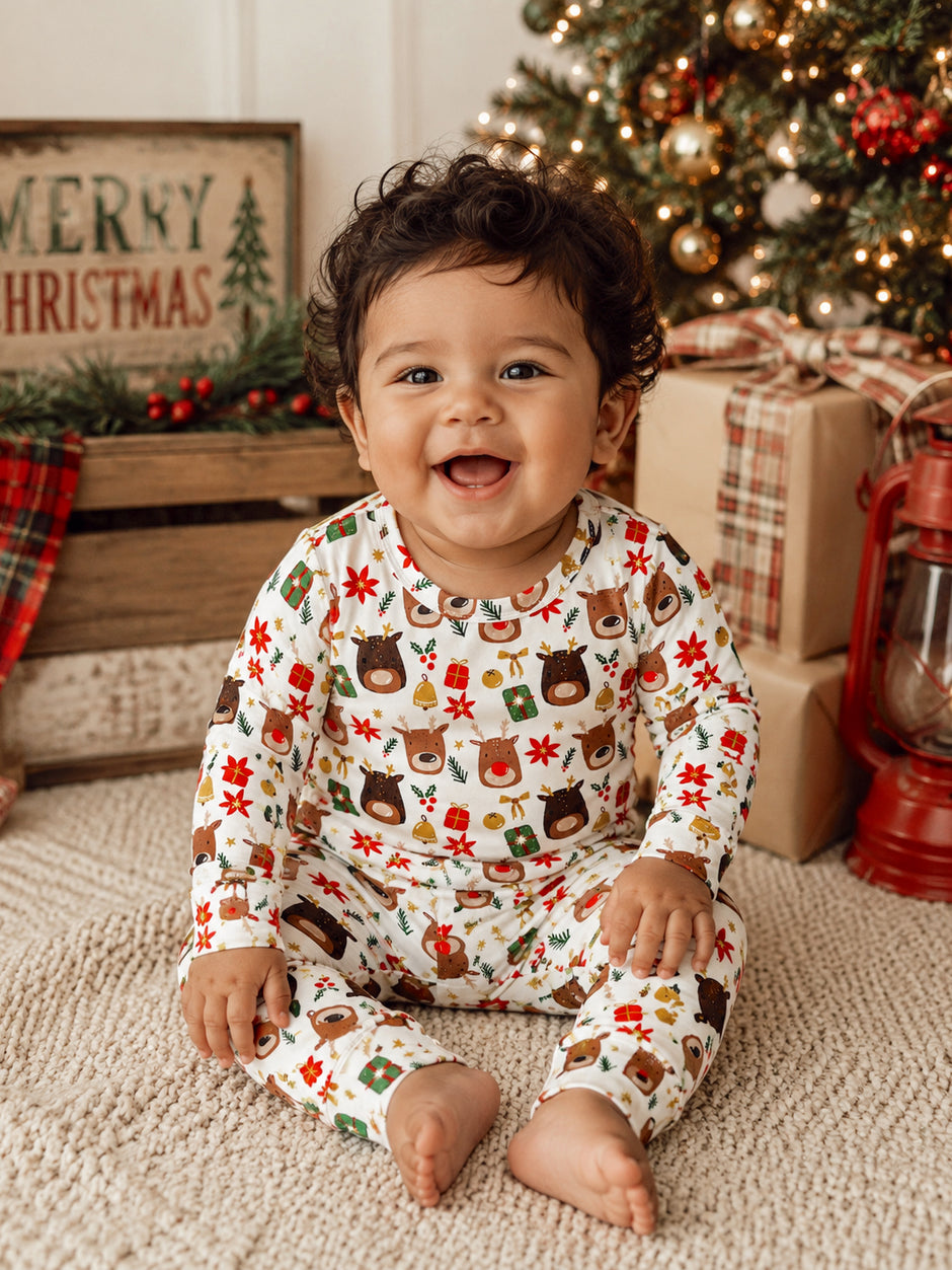 Smiling baby in festive clothing sitting on a rug near a Christmas tree and holiday decorations.
