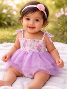 Smiling baby in a floral swimsuit with a purple tulle skirt and a pink headband, sitting on a blanket outdoors.