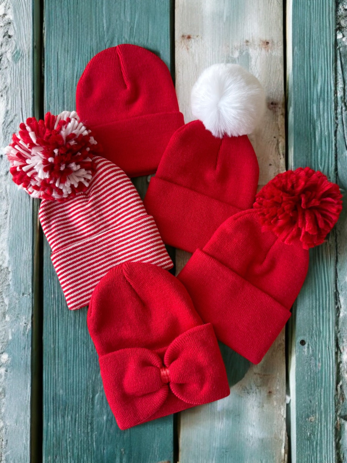 Five red beanies, including pom-pom styles and a bow design, arranged on a wooden surface.