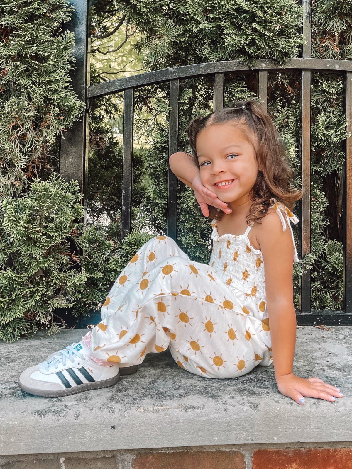 Smiling girl in a sun-printed outfit sitting on a stone wall with greenery in the background.