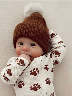 Baby wearing a brown knit hat and paw-print sweater, smiling and sucking on their hand.
