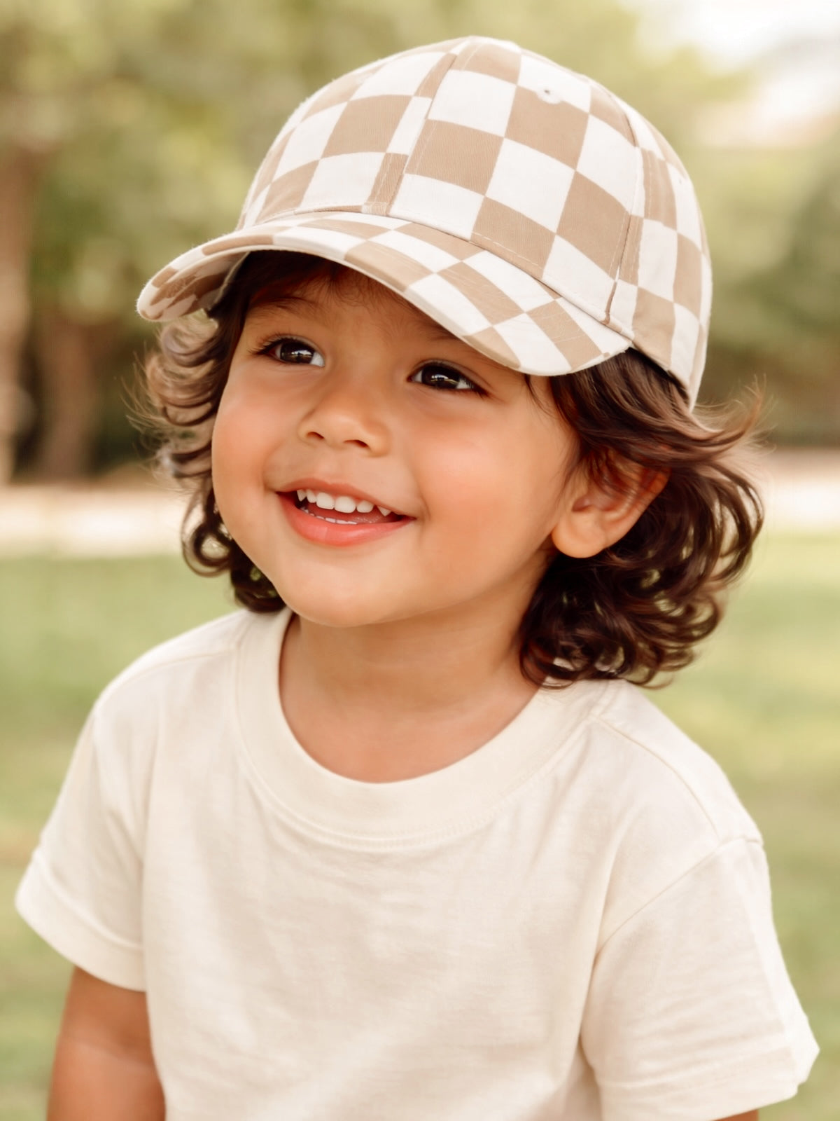 Smiling child with curly hair wearing a tan and white checkered cap, set outdoors against a leafy background.