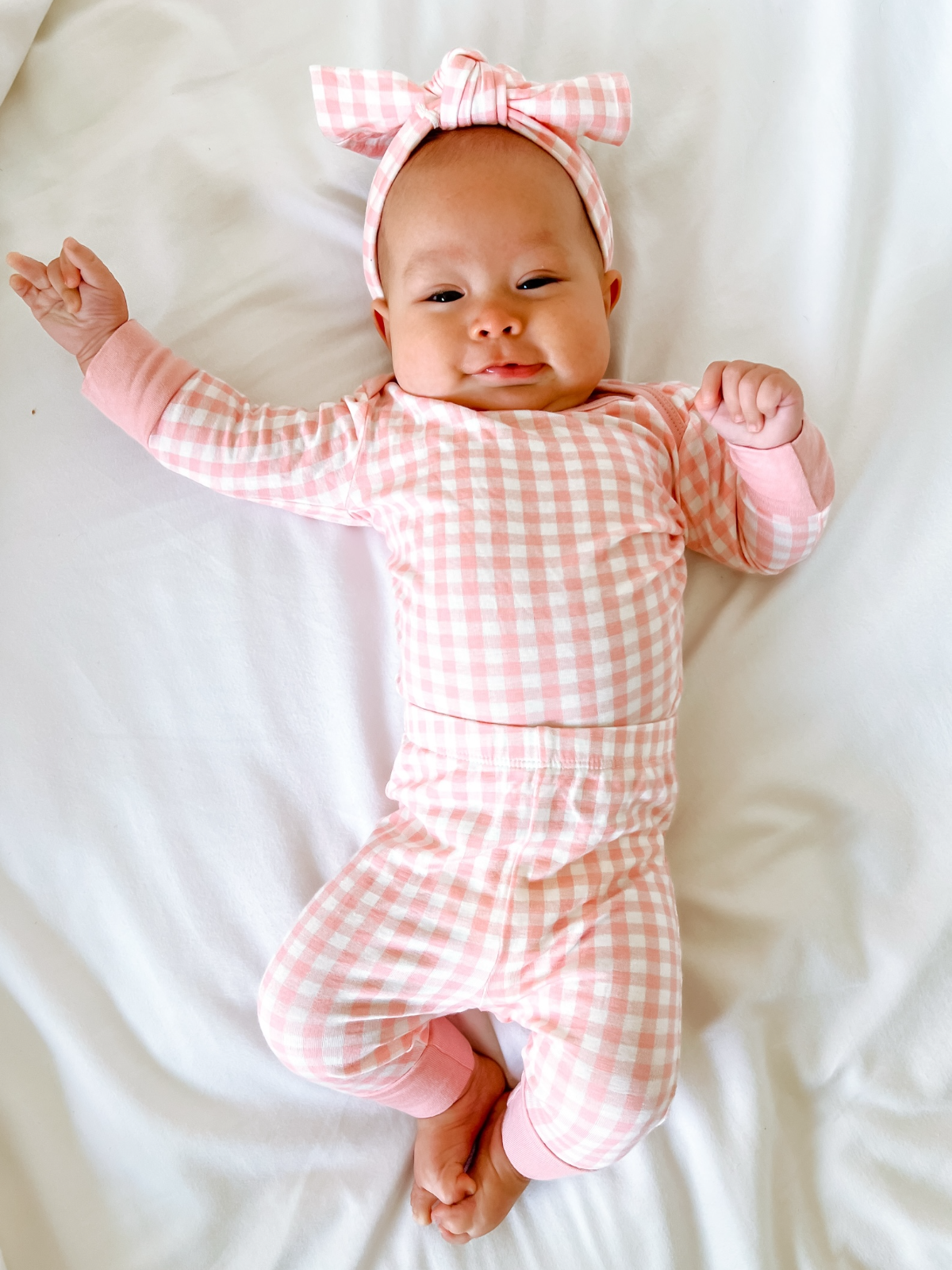 Smiling baby in pink gingham outfit and headband lying on a white blanket.