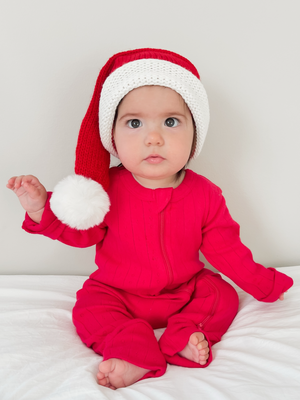 Baby in a red outfit and Santa hat, sitting on a white bed, looking curiously at the camera.