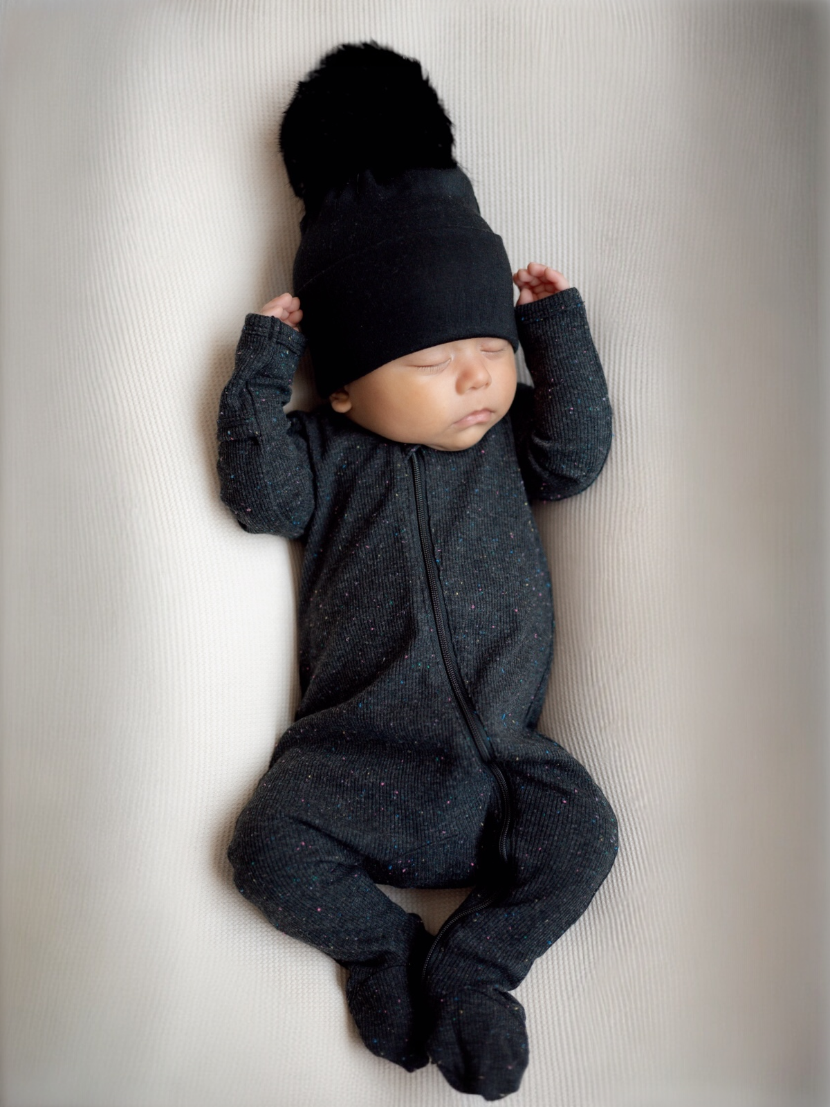 Baby sleeping peacefully in a cozy black outfit and beanie with a pom-pom, resting on a soft white surface.