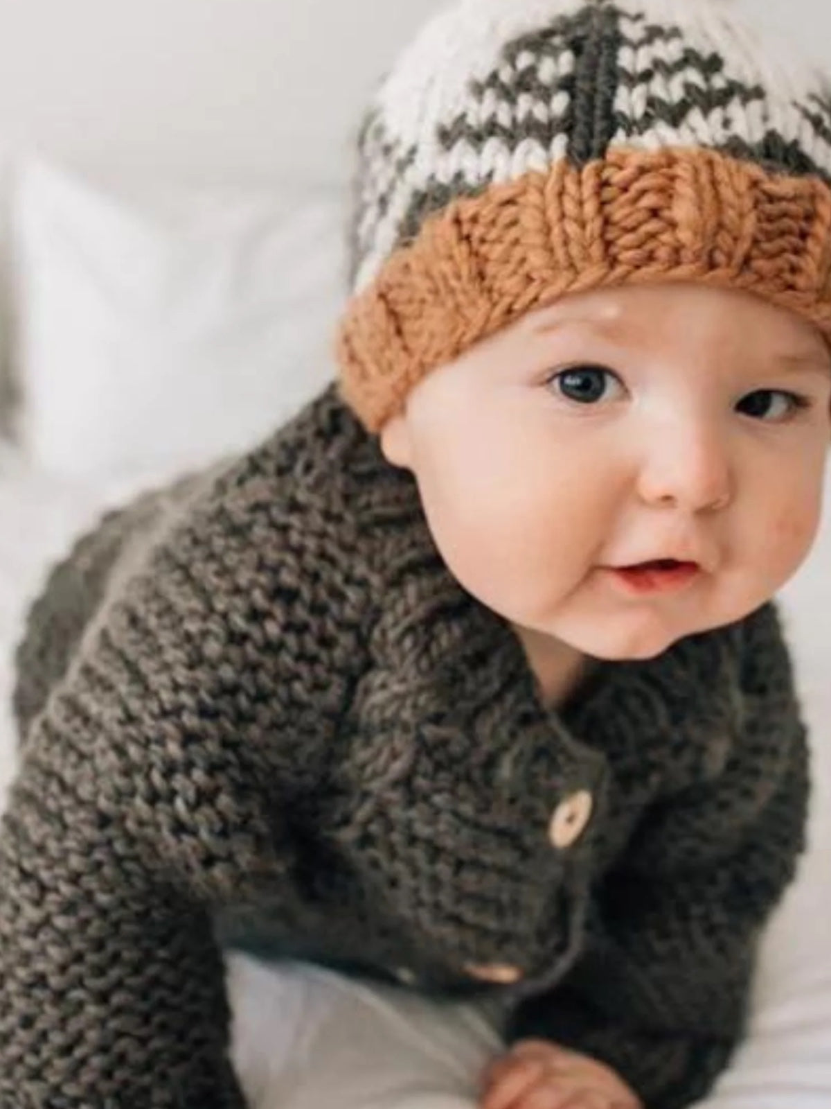 Baby wearing a knitted sweater and hat, smiling while crawling on a bed with white sheets.