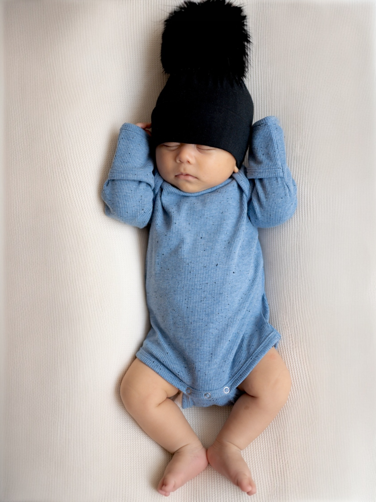 Baby wearing a blue onesie and black hat with pom-pom, sleeping peacefully on a textured surface.