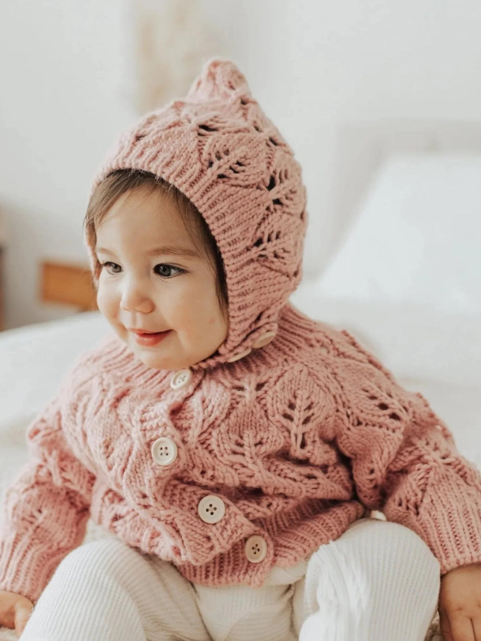 Smiling toddler in a pink knitted sweater and matching hat, sitting on a bed with a cozy backdrop.