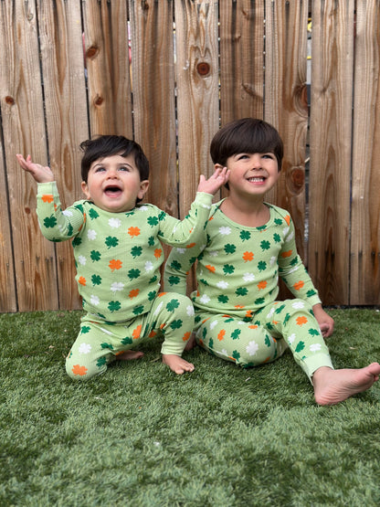 Two children in matching green pajama sets with shamrock patterns, sitting on grass and smiling joyfully.