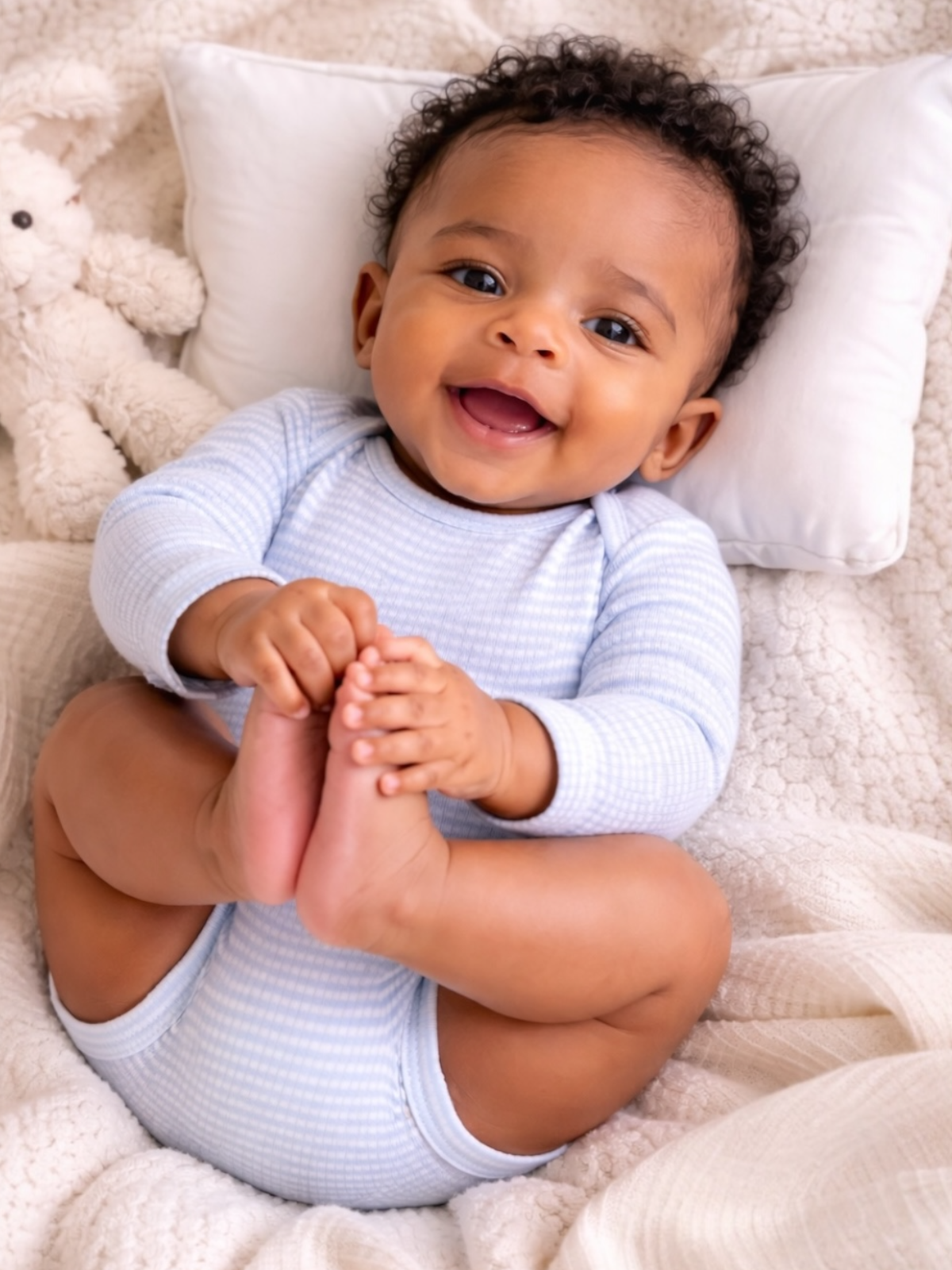 Smiling baby in light blue outfit, playfully holding feet, surrounded by soft blankets and a plush toy.