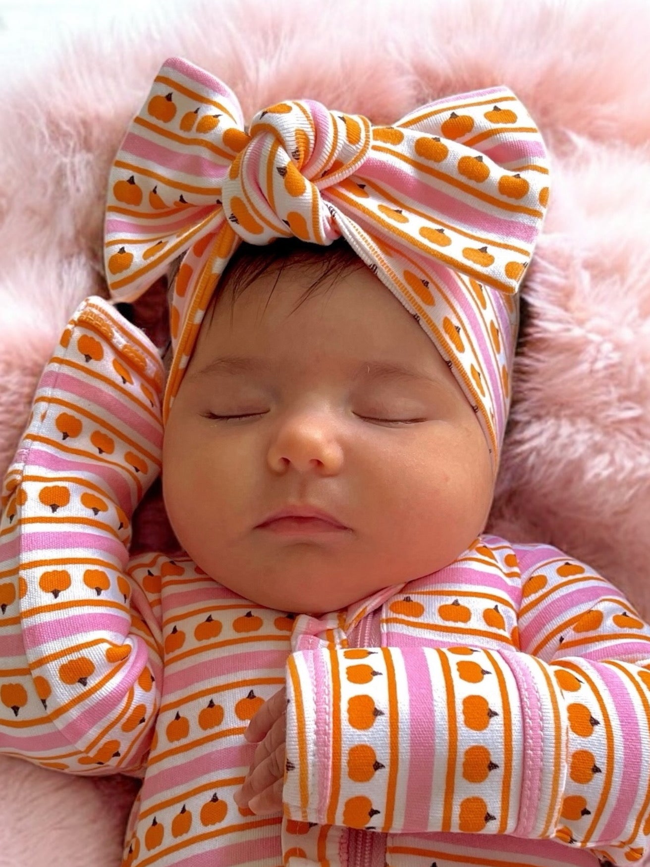 Infant girl asleep with a patterned headband and pajamas on a soft pink blanket.
