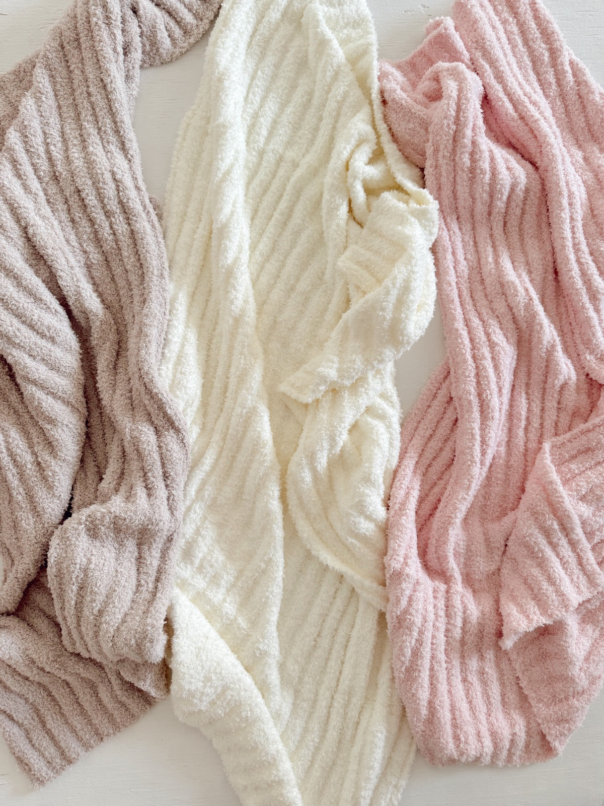 Three soft, textured blankets in brown, white, and pink, arranged casually on a light surface.