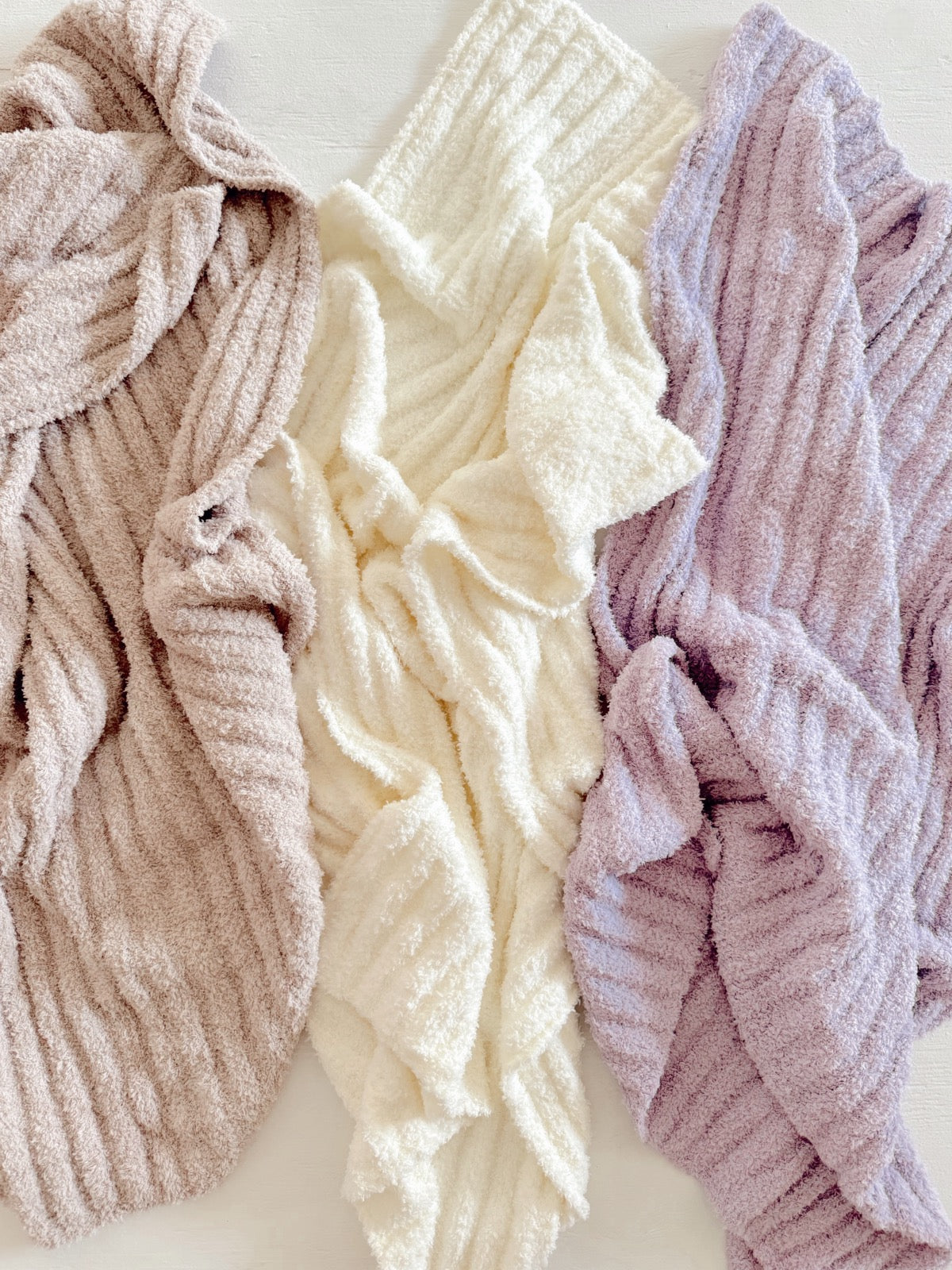 Three cozy, textured blankets in taupe, cream, and lavender, layered on a light surface.
