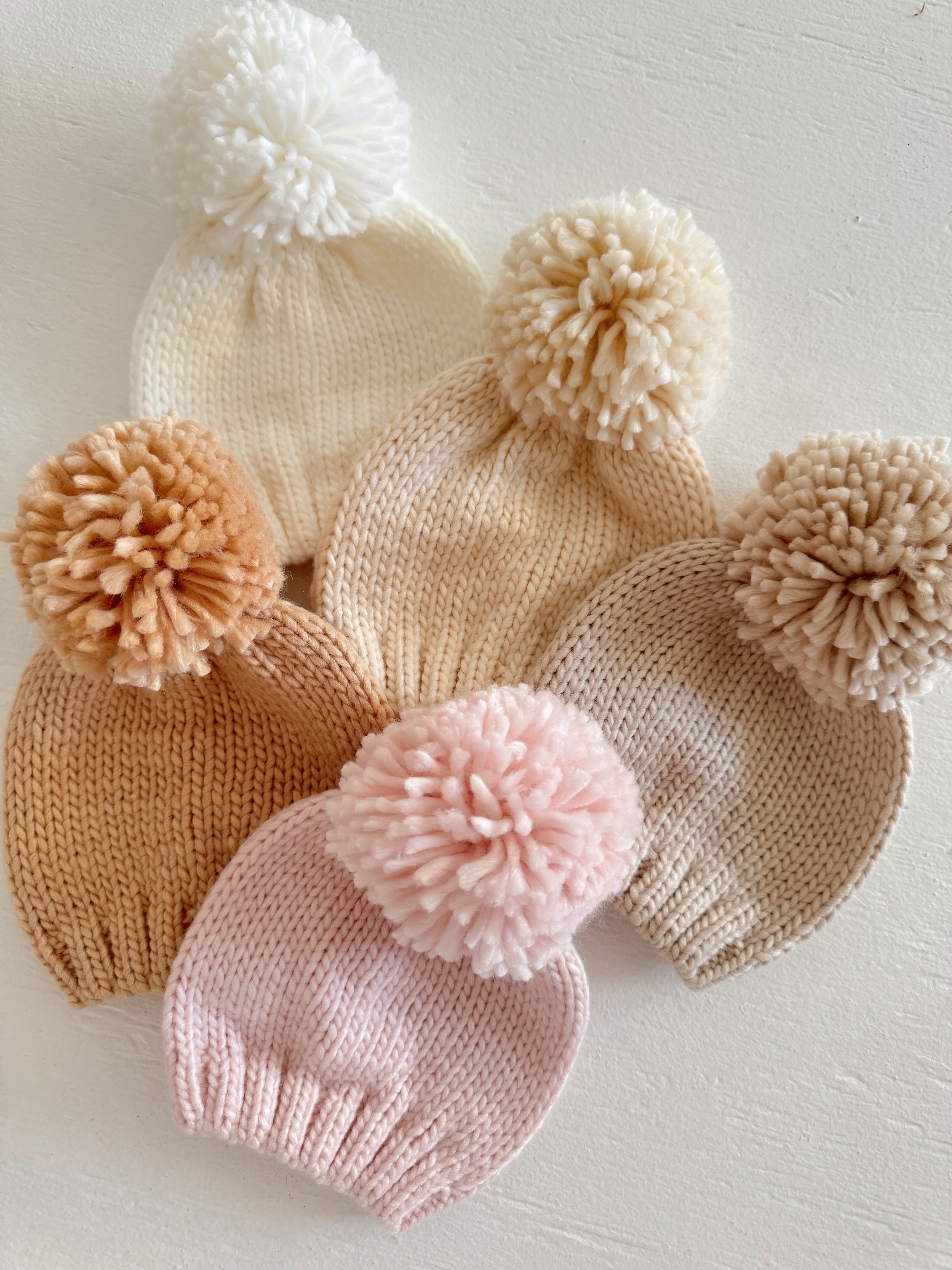 Set of six knitted beanies in neutral tones, each adorned with a fluffy pom-pom on top.