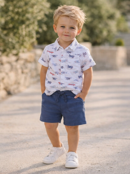 Smiling young boy in a patterned shirt and shorts standing outdoors.