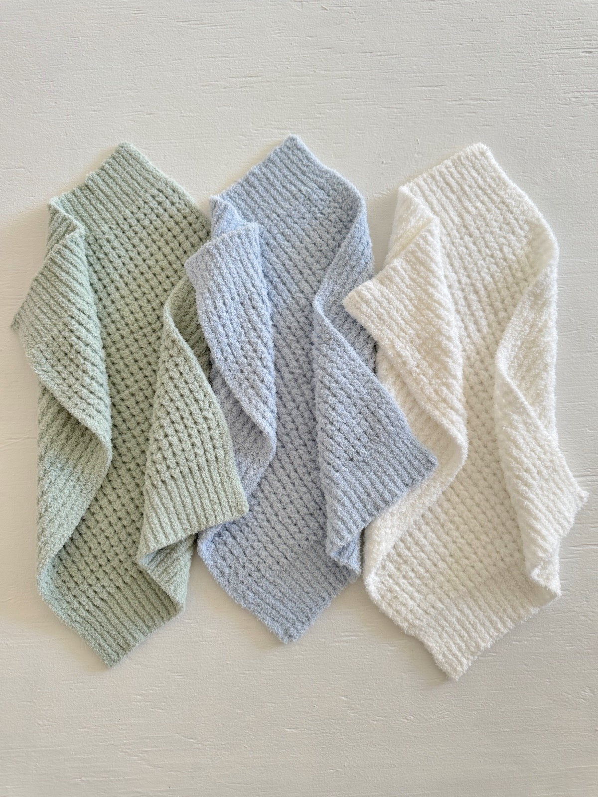 Three textured towels in soft colors: green, blue, and white, arranged on a light surface.