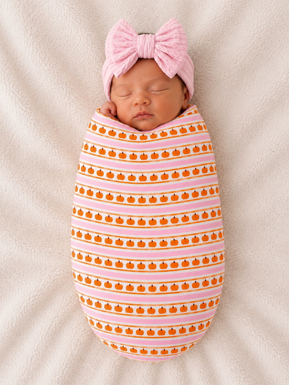 Newborn wrapped in pumpkin-patterned fabric, wearing a pink bow, resting on a soft, light-colored blanket.