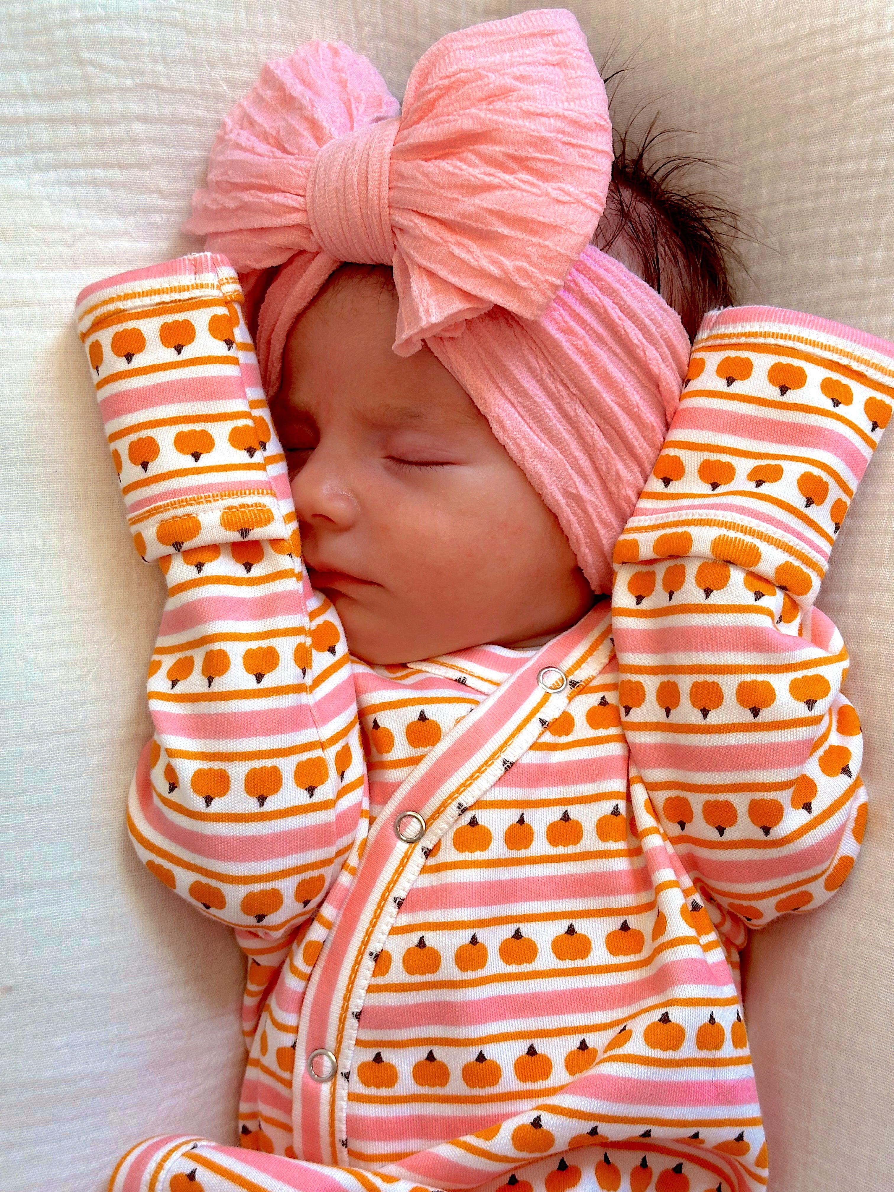 Baby sleeping peacefully in striped pumpkin-patterned outfit with a large pink bow headband.