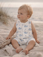 Child sitting on the sand in whale-patterned outfit, looking towards the ocean with gentle waves in the background.