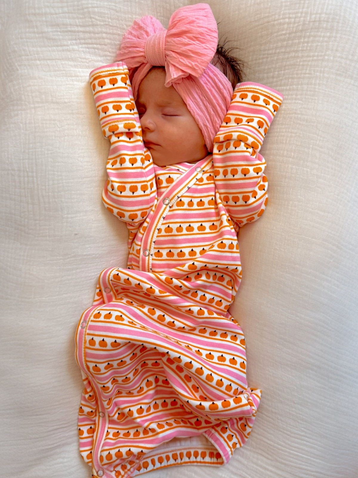 Sleeping newborn in a pumpkin-patterned outfit with a pink headband, against a soft white background.