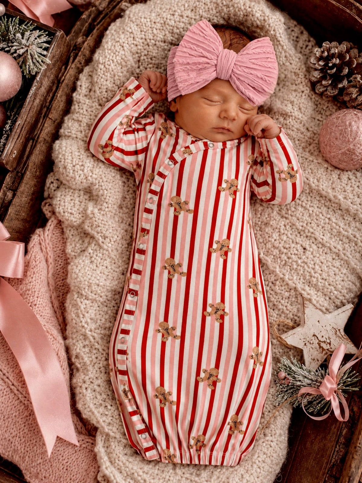 Newborn in red and white striped outfit with gingerbread prints, wearing a pink bow, resting on a cozy blanket.