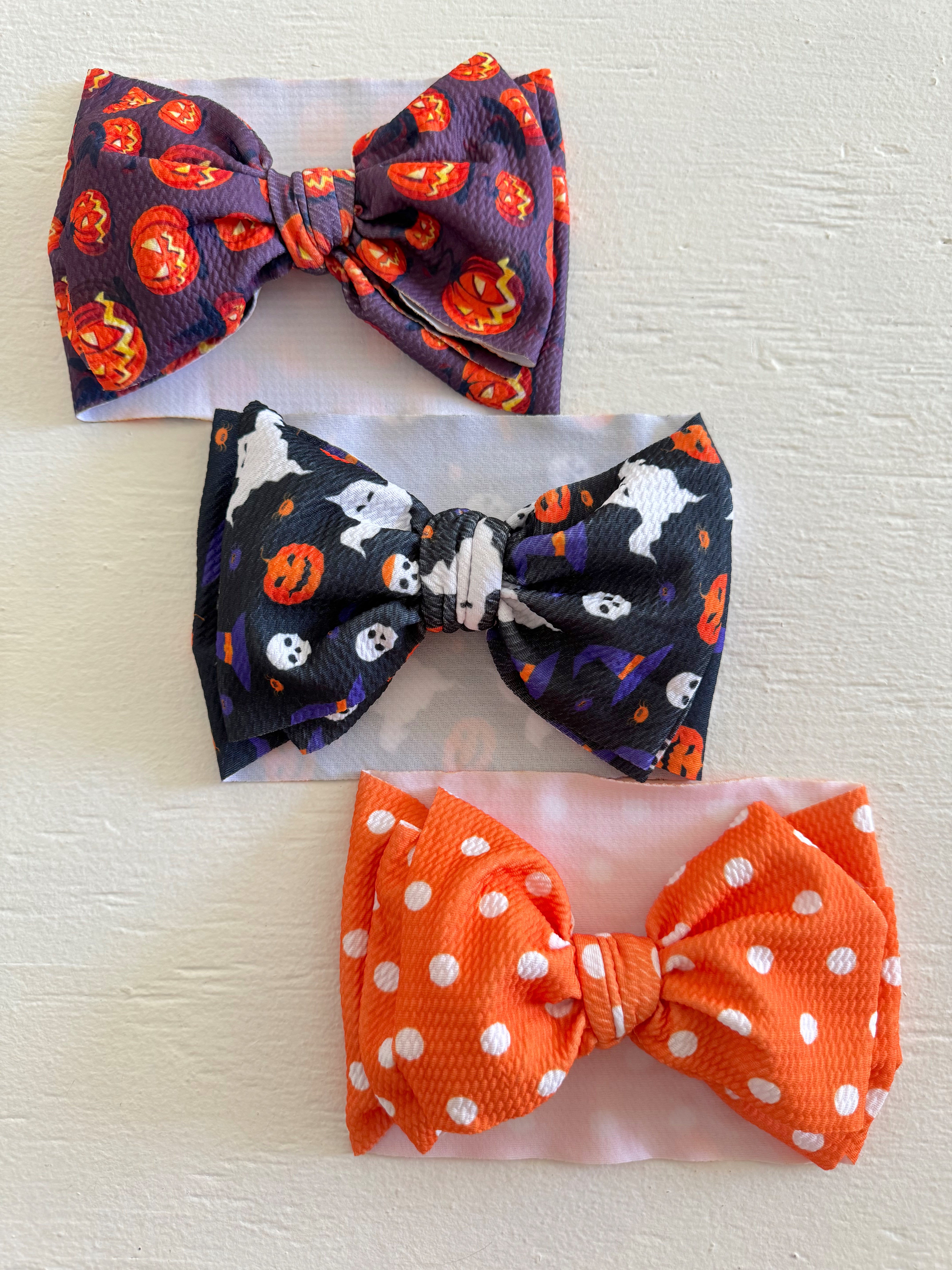 Three colorful hair bows: purple with pumpkins, black with ghosts, and orange with white polka dots.
