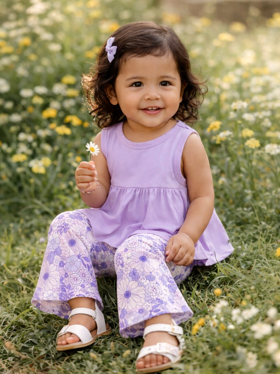 Smiling girl in a floral field, wearing a purple top and patterned pants, holding a daisy flower.