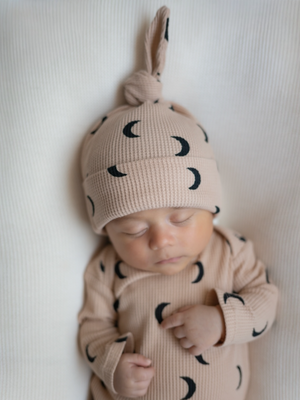 Sleeping baby wearing a soft, moon-patterned outfit and hat, lying on a textured white background.