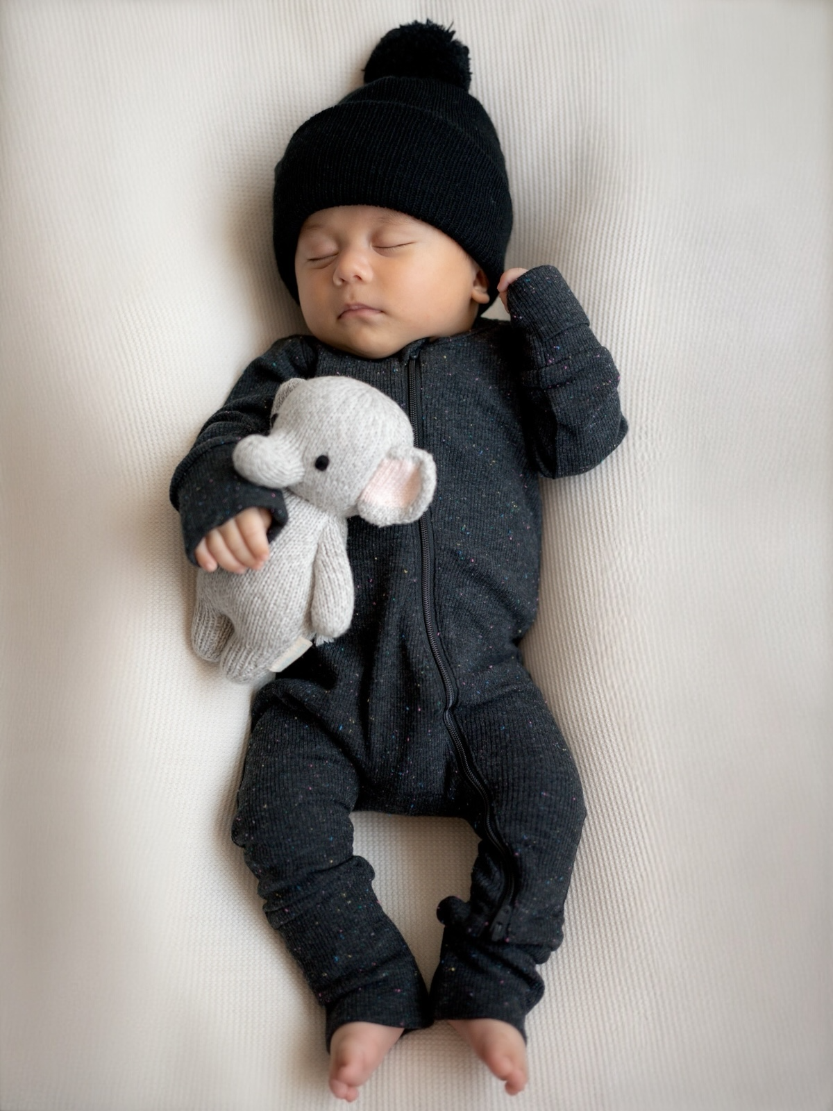 Sleeping baby in a black knit hat, wearing a dark onesie and holding a plush elephant toy.