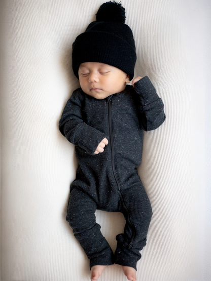 Baby in a black zip-up onesie and pom-pom hat sleeping peacefully on a light-colored surface.