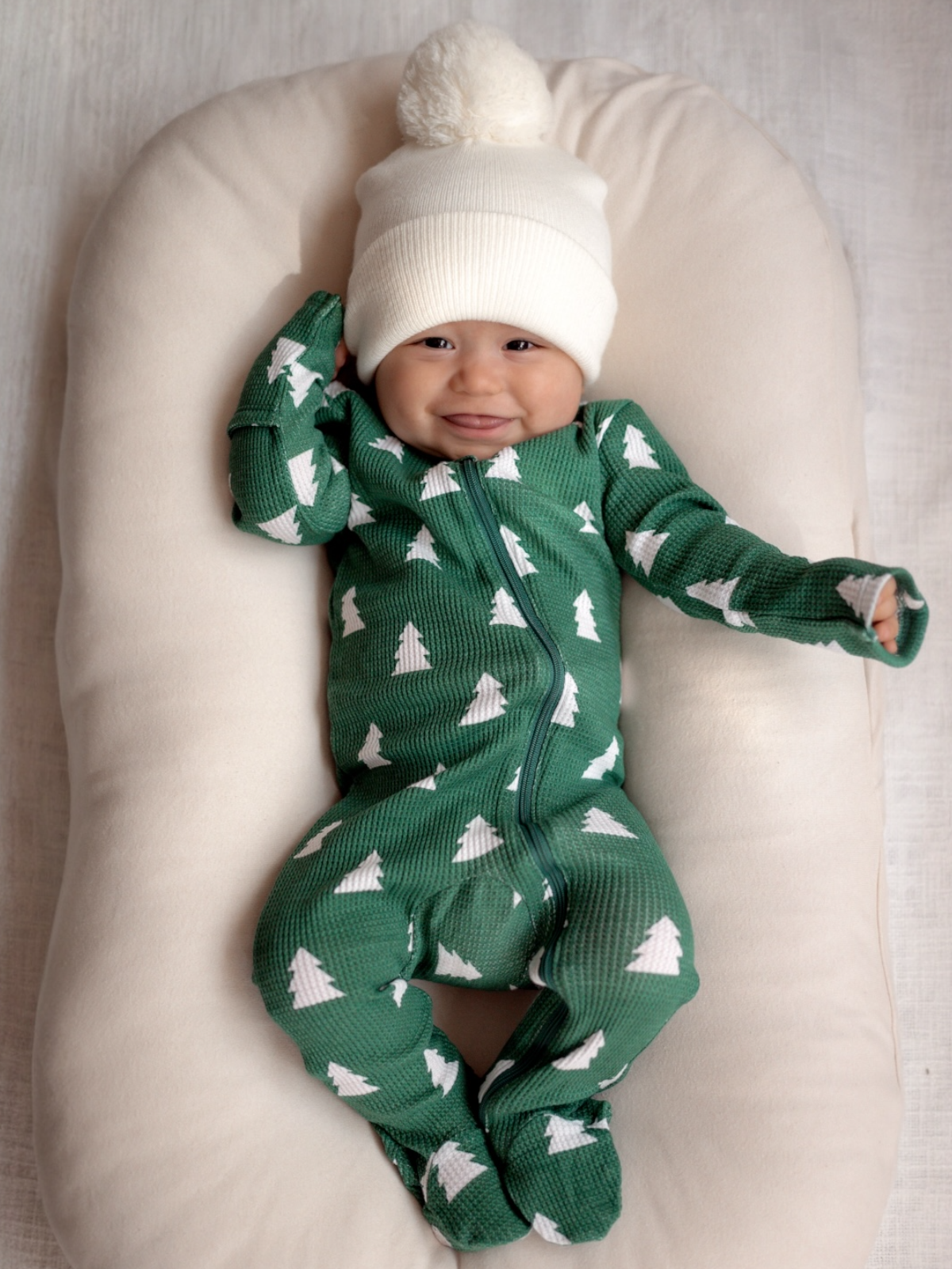 Smiling baby in a green onesie with trees, wearing a white pom-pom hat, resting on a soft pillow.