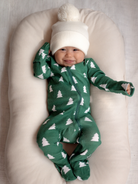 Smiling baby in a green onesie with trees, wearing a white pom-pom hat, resting on a soft pillow.