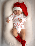 Baby in festive red hat and candy cane onesie, nestled on a cozy surface, looking curiously at the camera.