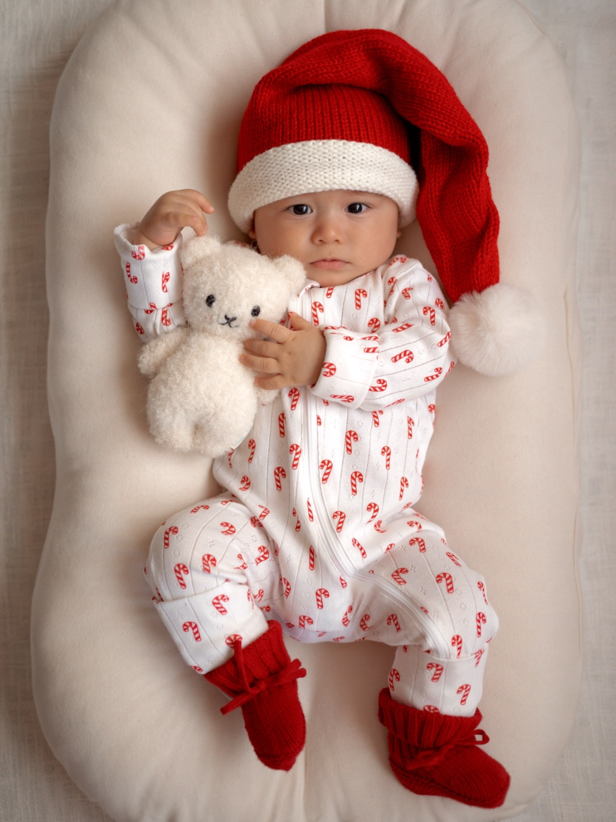 Baby in a red and white candy cane onesie, wearing a Santa hat, holding a teddy bear, cozy in a plush setting.